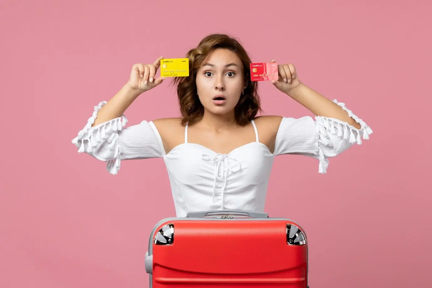 Вид спереди: молодая женщина с сумкой для отпуска держит банковские карты на фоне розовой стены.